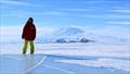 Catherine Walker stands on the McMurdo Ice Shelf in Antarctica in Oct 2014. In the background, U.S. Antarctic Program's McMurdo Station is visible at the base of Mt. Erebus, an active volcano, along with a C-130 aircraft delivering people & cargo